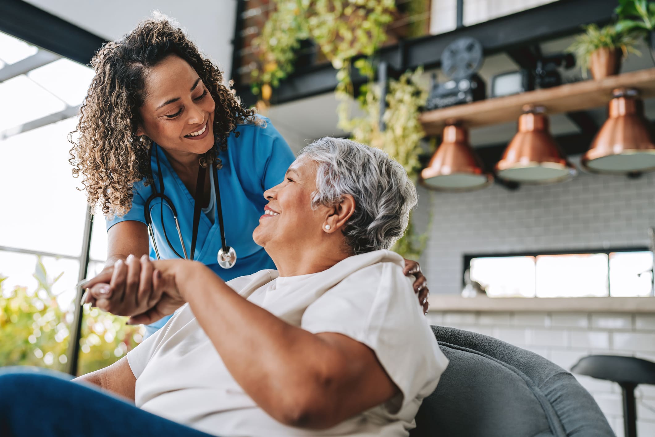 A young woman working as a home health aide in the allied health industry smiles down at an elderly woman with gray hair and holds her hand.